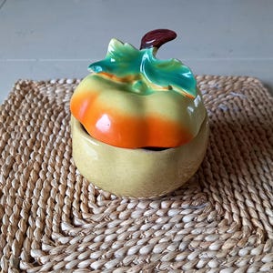 Può includere: Un contenitore in ceramica a forma di zucca con coperchio. Il coperchio è una zucca con foglie verdi e un gambo marrone. Il contenitore è giallo con sfumature arancioni e gialle. Il contenitore è su un tappetino intrecciato.