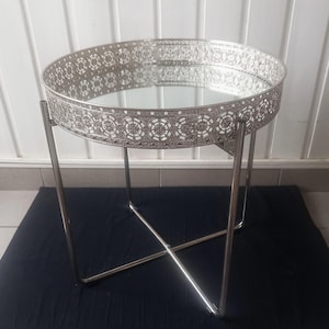 Puede incluir: Una mesa auxiliar redonda de color plateado con una tapa de espejo y bordes decorativos de filigrana. La mesa tiene un diseño geométrico y está sostenida por cuatro patas delgadas e inclinadas. La mesa está sobre una superficie negra.