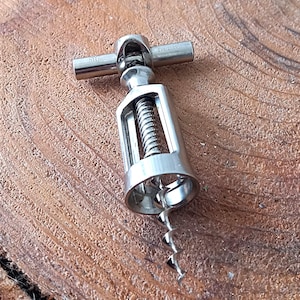 Può includere: Un cavatappi argentato con una spirale e un manico, progettato per rimuovere i tappi dalle bottiglie di vino. Lo strumento in metallo è mostrato su una superficie di legno. Il cavatappi è in metallo.
