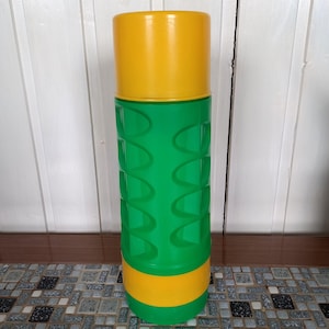 Può includere: Un contenitore cilindrico alto con parte superiore e inferiore gialle e un corpo verde strutturato. La sezione verde presenta un motivo ripetuto di rientranze curve. Il contenitore è in plastica e sembra un thermos o una borraccia.