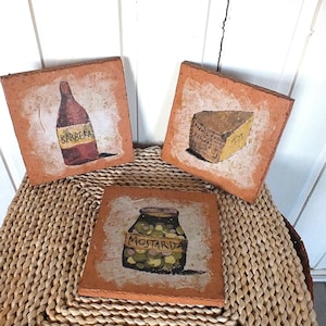 Könnte beinhalten: Drei quadratische Untersetzer in Terrakotta-Farbe mit handgemalten Bildern. Einer zeigt eine Weinflasche mit der Aufschrift "Barbera", ein weiterer ein Käsekeil und der dritte ein Glas "Mostarda". Die Untersetzer liegen auf einer gewebten Oberfläche.