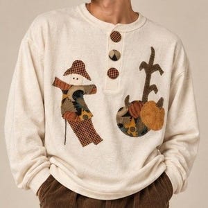 May include: A cream-coloured long-sleeve henley shirt featuring three round buttons and fabric appliqués. The design includes a scarecrow figure in a plaid hat and a separate patch depicting a pumpkin and corn stalk in shades of orange, brown, and black.