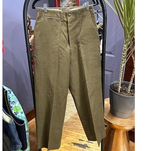 May include: Olive green trousers hanging on a black metal rack. The pants have a classic, straight-leg design with a button and zipper closure. The fabric appears to be a durable, woven material. The trousers are displayed in a well-lit setting.