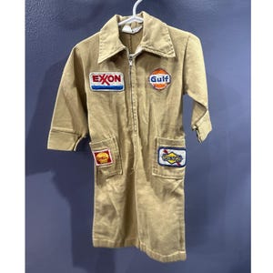 May include: Tan children's mechanic jumpsuit with a zipper closure and collared neckline. The jumpsuit features embroidered patches for Exxon, Gulf, Shell, and Sunoco. The sleeves are short, and there are pockets on the chest and lower front.