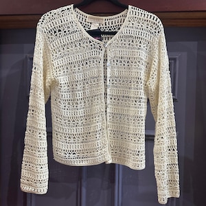 May include: Cream-colored, open-weave crochet cardigan with long sleeves and a V-neck. The button-up sweater is made of a light, airy material and hangs on a black hanger against a purple background. The cardigan has a delicate, textured appearance.