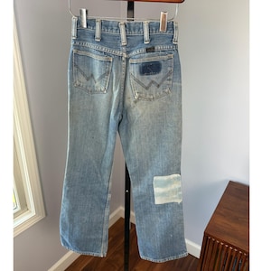 May include: Faded light blue denim jeans with a patch on the right leg. The jeans feature a classic five-pocket design, a zip fly, and a Wrangler patch on the back pocket. The jeans are suspended from a wooden clothes hanger.