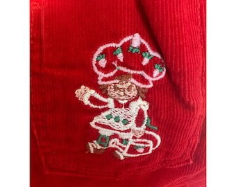 Op de afbeelding: Rode corduroy stof met een borduursel van het Strawberry Shortcake-personage op een zak. Het personage draagt een witte jurk met groene stippen en een aardbeienhoed. De zak zit op een groter stuk rode stof.