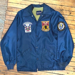 Puede incluir: Chaqueta Duckster azul marino con cierre de botones, cuello y múltiples parches. Los parches incluyen texto y emblemas, como "USS Forrestal" y "United States Ship Ajax AR-6". La chaqueta tiene forro verde y puños elásticos.