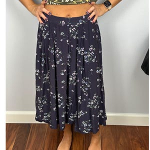 May include: A navy blue midi skirt with a floral pattern in white, light blue, and pink. The skirt has a pleated waist and falls to mid-calf length. The fabric appears to be lightweight and flowing, suitable for warmer weather.