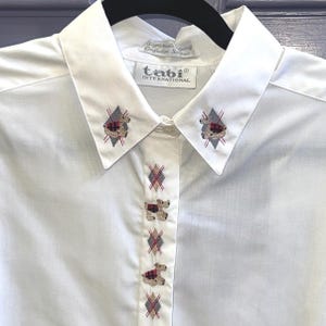 May include: White button-down shirt with embroidered teddy bear and argyle pattern details. The shirt features a pointed collar and the "tabi INTERNATIONAL" label. The embroidery includes red, gray, and brown accents.