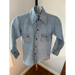 Puede incluir: Camisa abotonada azul claro con mangas largas. Cuenta con dos bolsillos en el pecho y botones negros. La etiqueta de la marca Levi's es visible en el cuello. Una etiqueta naranja de Levi's está en el bolsillo. La camisa está colgada en una percha.