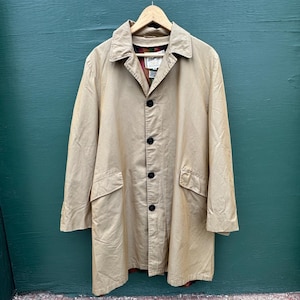May include: A tan, button-down coat with a collared neckline and two front pockets. The coat is hanging on a wooden hanger against a teal-colored background. The coat has a classic, timeless design.