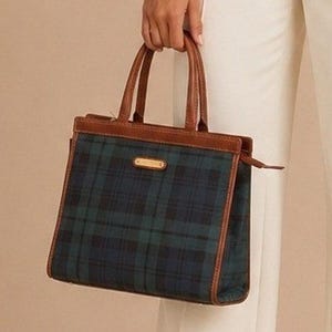 May include: A structured handbag featuring a dark green and navy blue plaid pattern. The bag is accented with brown leather trim along the top, base, and handles. A small rectangular gold-toned metal plaque is centered on the front upper panel.