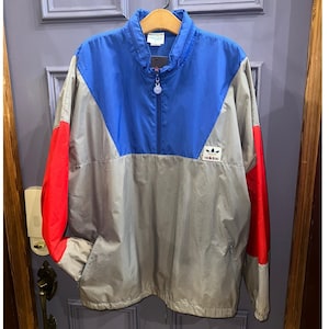 Op de afbeelding: Een Adidas-trainingsjack met kleurblokken in blauw, grijs en rood. De jas heeft een opstaande kraag, een halve ritssluiting en het Adidas-logo op de borst. De mouwen zijn grijs met rode accenten en de body is grijs en blauw.