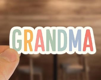 May include: A colorful sticker with the word "GRANDMA" in pastel colors. The letters are outlined in white, with a light green "G", orange "R", pink "A", light blue "N", light blue "D", pink "M", and red "A".