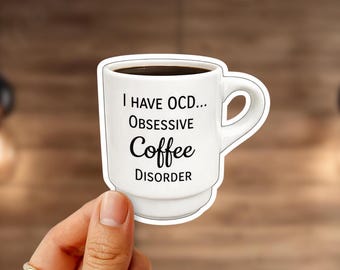 May include: White coffee mug sticker with the text "I HAVE OCD... OBSESSIVE Coffee DISORDER". The mug is filled with dark coffee. The sticker is held by a hand with a gold ring.