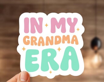 May include: A white sticker with the text "IN MY GRANDMA ERA" in pink, orange, and teal lettering. The sticker has a rounded shape and is decorated with small star accents. The sticker is held by a hand.