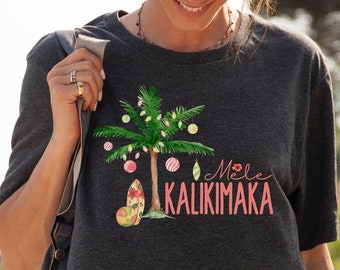 May include: Dark gray t-shirt featuring a tropical Christmas tree design with ornaments and a surfboard. The text "Mele Kalikimaka" is in pink. The shirt is made of a soft material.