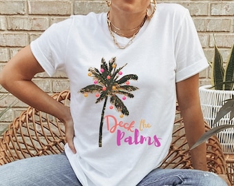 May include: White t-shirt with a Christmas palm tree design, adorned with baubles. The text "Deck the Palms" is in pink and orange. A casual, festive top, perfect for the holiday season.