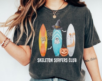 May include: A charcoal grey t-shirt featuring a graphic of three surfboards, a pumpkin, and a ghost. The centre surfboard has a witch hat, and the text SKELETON SURFERS CLUB is printed in white below the surfboards with a spiderweb and bats.