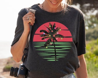 May include: Dark gray t-shirt featuring a palm tree decorated with Christmas lights and ornaments against a red and green sunset design. The shirt is made of a soft material and is suitable for casual wear.