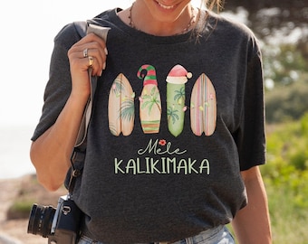 May include: A dark grey t-shirt with a festive design of surfboards decorated with Christmas elements. The shirt has the words "Mele Kalikimaka" in green, with a small flower accent. The design includes palm trees and Santa hats.