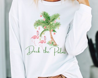 May include: White long-sleeve shirt featuring a watercolor design of a decorated palm tree and two pink flamingos. The text "Deck the Palms" is written in green script below the image. The shirt is made of a soft material.