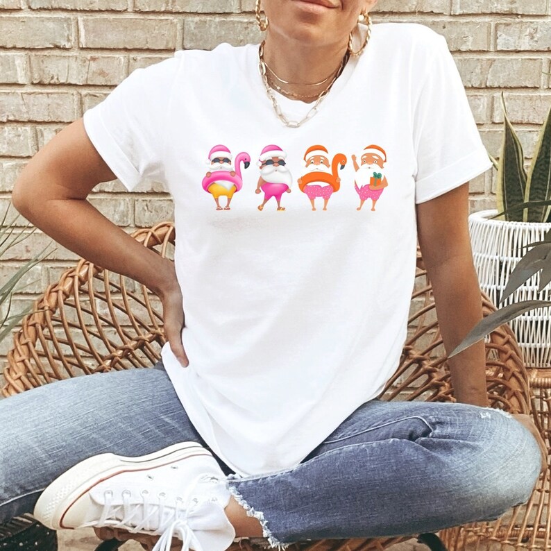 May include: White t-shirt with a playful design of four Santa Claus figures, each with a flamingo theme. The Santas wear sunglasses and swim shorts. The design uses pink, yellow, and orange, creating a fun, summery feel. The t-shirt is a casual wear item.