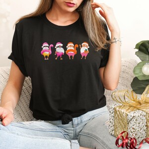 May include: Black t-shirt with a humorous Christmas design. It features Santa Claus figures with flamingo bodies, each wearing a pink hat and white beard. The Santas are in different poses, creating a fun, festive look.