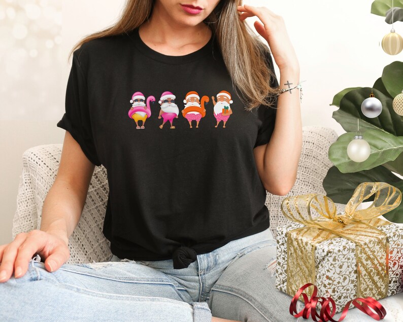 May include: Black t-shirt with a humorous Christmas design. It features Santa Claus figures with flamingo bodies, each wearing a pink hat and white beard. The Santas are in different poses, creating a fun, festive look.