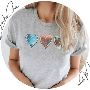 A gray t-shirt with three hearts printed on the front. Each heart has a different beach scene inside. The first heart has a blue ocean scene, the second heart has a sunset scene, and the third heart has a palm tree scene.