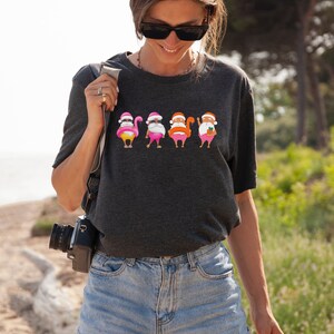May include: Dark grey t-shirt with a graphic of four Santas in summer outfits. Each Santa has a different accessory, including a flamingo float, sunglasses, and a cocktail. The Santas are wearing pink shorts and Santa hats.