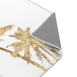 A white area rug with a design of three palm trees in shades of brown and yellow. The rug has a gray backing and is stitched around the edges.