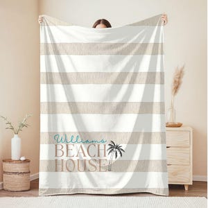May include: A large, striped beach blanket with alternating beige and white horizontal stripes. The blanket features the text "Williams BEACH HOUSE" in teal and brown, with a palm tree graphic. The blanket is held up by a person.