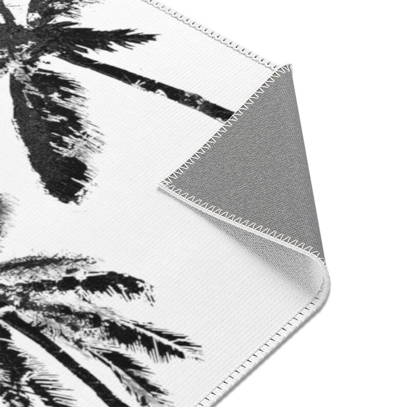 Peut inclure: Un tapis blanc avec un motif de palmier noir et blanc. Le tapis a un dos gris et est cousu sur les bords.