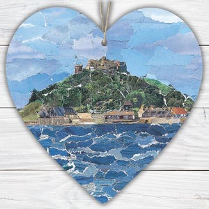 May include: Heart-shaped wooden wall hanging featuring a collage of a coastal landscape. The artwork depicts a castle atop a green hill, buildings, and blue water. A jute string is attached for hanging. The background is a white, wooden surface.