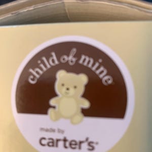 May include: A brown circle with the text 'child of mine' in white text. Inside the circle is a cartoon image of a light brown teddy bear. Below the circle is the text 'made by carter's' in white text.
