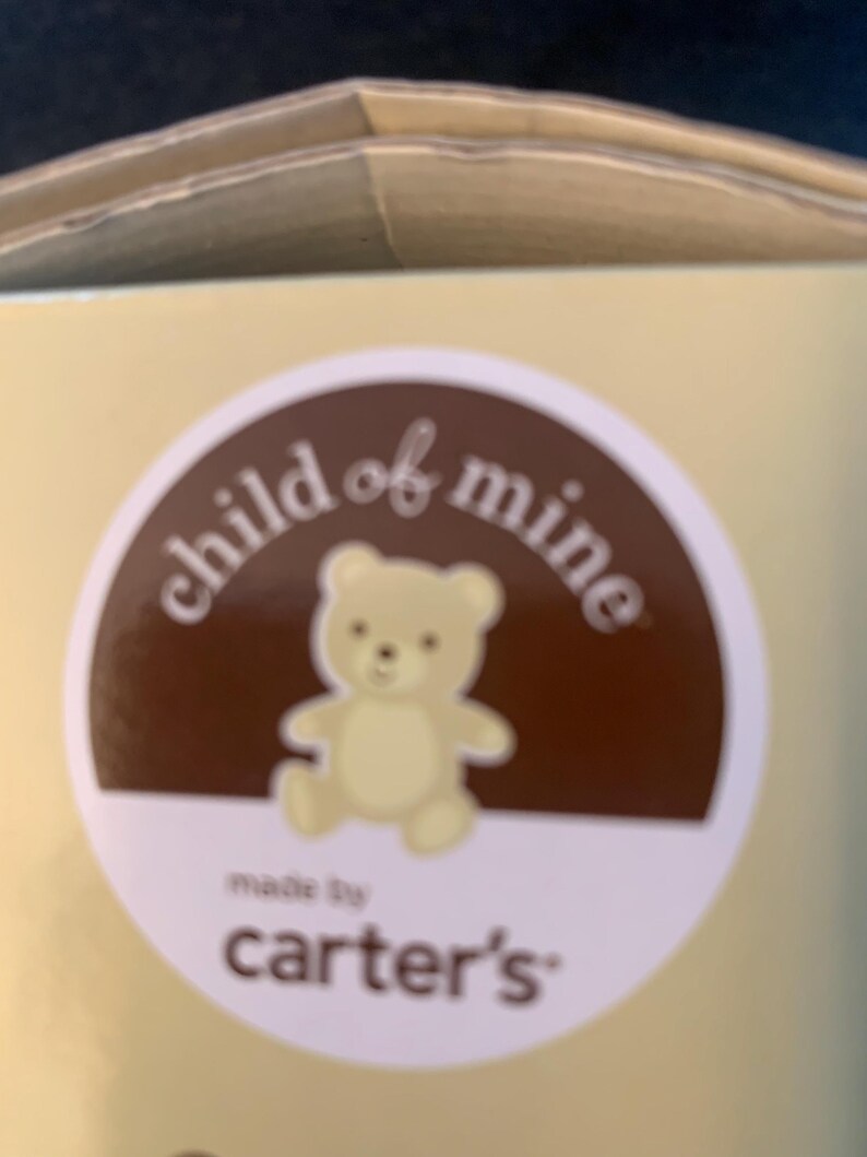 May include: A brown circle with the text 'child of mine' in white text. Inside the circle is a cartoon image of a light brown teddy bear. Below the circle is the text 'made by carter's' in white text.
