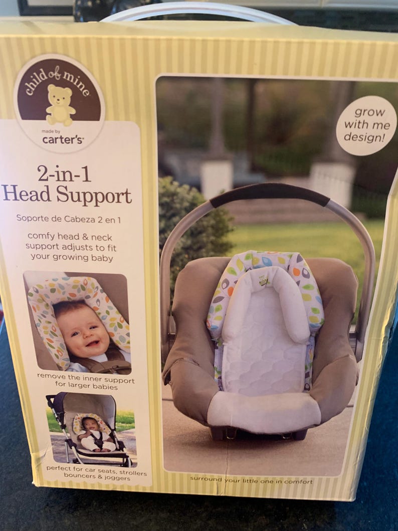 May include: A box for a 2-in-1 baby head support. The box is yellow with brown stripes and features a cartoon baby bear. The product is made by Carter's and is designed to grow with the baby. The box shows a baby in a car seat using the head support. The text on the box reads 'child of mine', 'made by carter's', '2-in-1 Head Support', 'Soporte de Cabeza 2 en 1', 'comfy head & neck support adjusts to fit your growing baby', 'remove the inner support for larger babies', 'perfect for car seats, strollers, bouncers & joggers', 'grow with me design!' and 'surround your little one in comfort'.