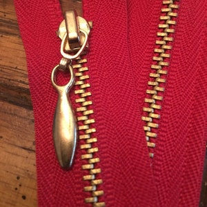 May include: Red zipper tape with a gold metal zipper pull and teeth. The zipper is closed.