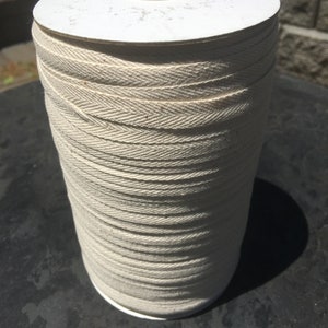 May include: A spool of off-white cotton twill tape. The tape is wound tightly around a white cardboard core.