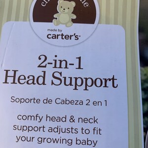 May include: A white and brown package for a 2-in-1 head support for babies. The package has a brown circle with the words 'child of mine' and 'made by carter's' in white text. The package also has the text '2-in-1 Head Support' in brown text and 'Soporte de Cabeza 2 en 1' in smaller brown text. The text 'comfy head & neck support adjusts to fit your growing baby' is also in brown text.