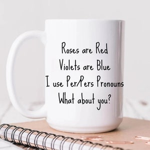 Puede incluir: Taza de cerámica blanca con texto negro que dice "Roses are Red, Violets are Blue, I use Per/Pers Pronouns, What about you?" La taza está sobre un cuaderno marrón con espiral dorado. La taza es de 15 oz.