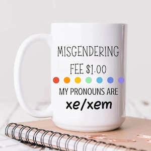 Könnte beinhalten: Weiße Keramik-Tasse mit dem Text "Misgendering Fee $1.00" und "My pronouns are xe/xem" in schwarzer Schrift. Die Tasse hat ein Regenbogen-Punkt-Design und fasst 444 ml Flüssigkeit. Die Tasse steht auf einem Notizbuch.