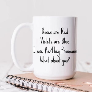 May include: White ceramic mug with a black handwritten message that reads "Roses are Red, Violets are Blue, I use He/They Pronouns, What about you?" The mug is sitting on a brown notebook with a black spiral binding. The mug is 15 fluid ounces.