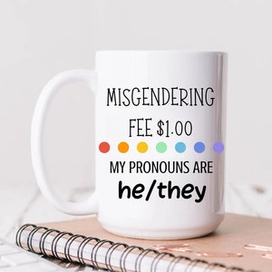 May include: White ceramic mug with the text "Misgendering Fee £1.00 My Pronouns Are he/they" in black text. The text is accompanied by a rainbow of six coloured dots. The mug is 15 oz.