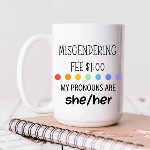May include: White ceramic mug with the text "Misgendering Fee $1.00 My Pronouns Are she/her" in black text. The text is surrounded by a rainbow of colored dots. The mug is 15 oz.