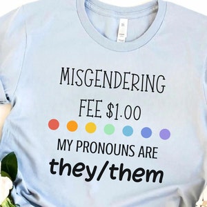 Könnte beinhalten: Hellblaues T-Shirt mit dem Text "Misgendering Fee $1.00" und einer Reihe von regenbogenfarbenen Punkten. Der Text "My Pronouns Are they/them" befindet sich unter den Punkten.