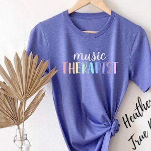 May include: A purple t-shirt with the text "music therapist" in rainbow colors. The text is in a playful, handwritten font.