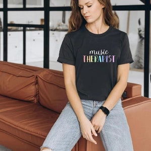 May include: A woman wearing a dark grey t-shirt with the text "music therapist" in rainbow colors. She is sitting on a brown leather couch.